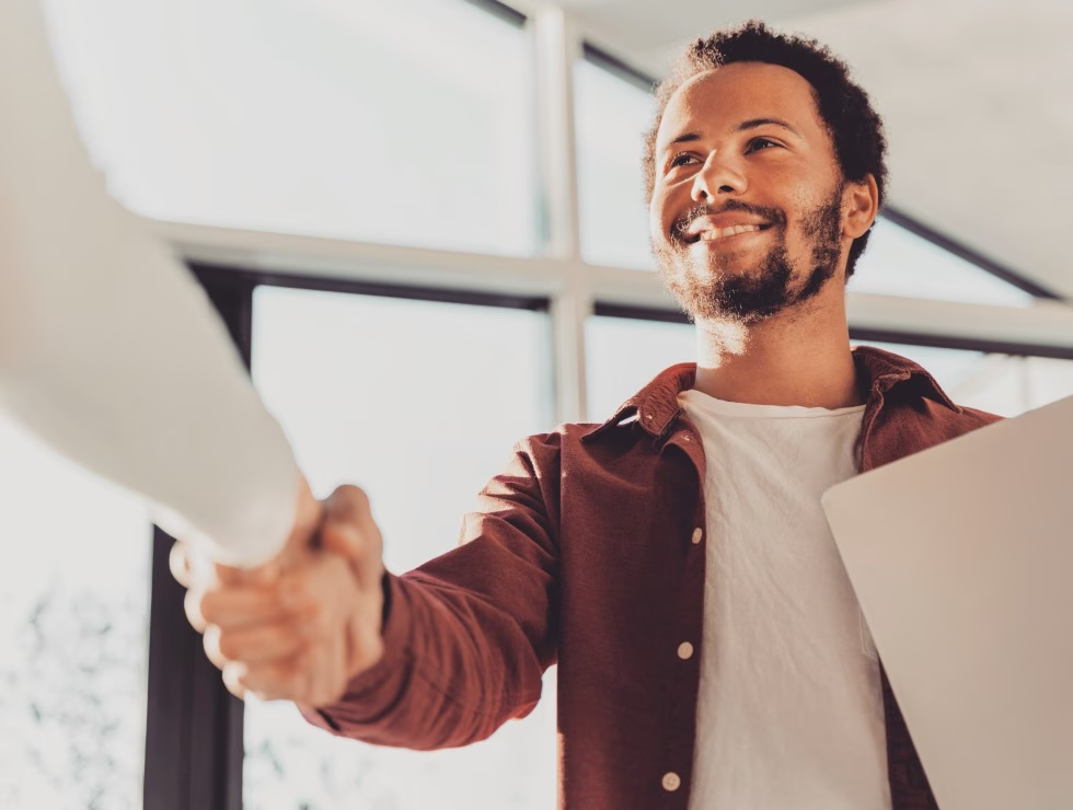 Jeune homme souriant serrant la main d’un interlocuteur dans un cadre professionnel lumineux